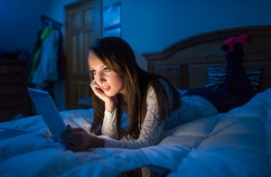Teenage girl in the dark lying on a bed working on a tablet computer.